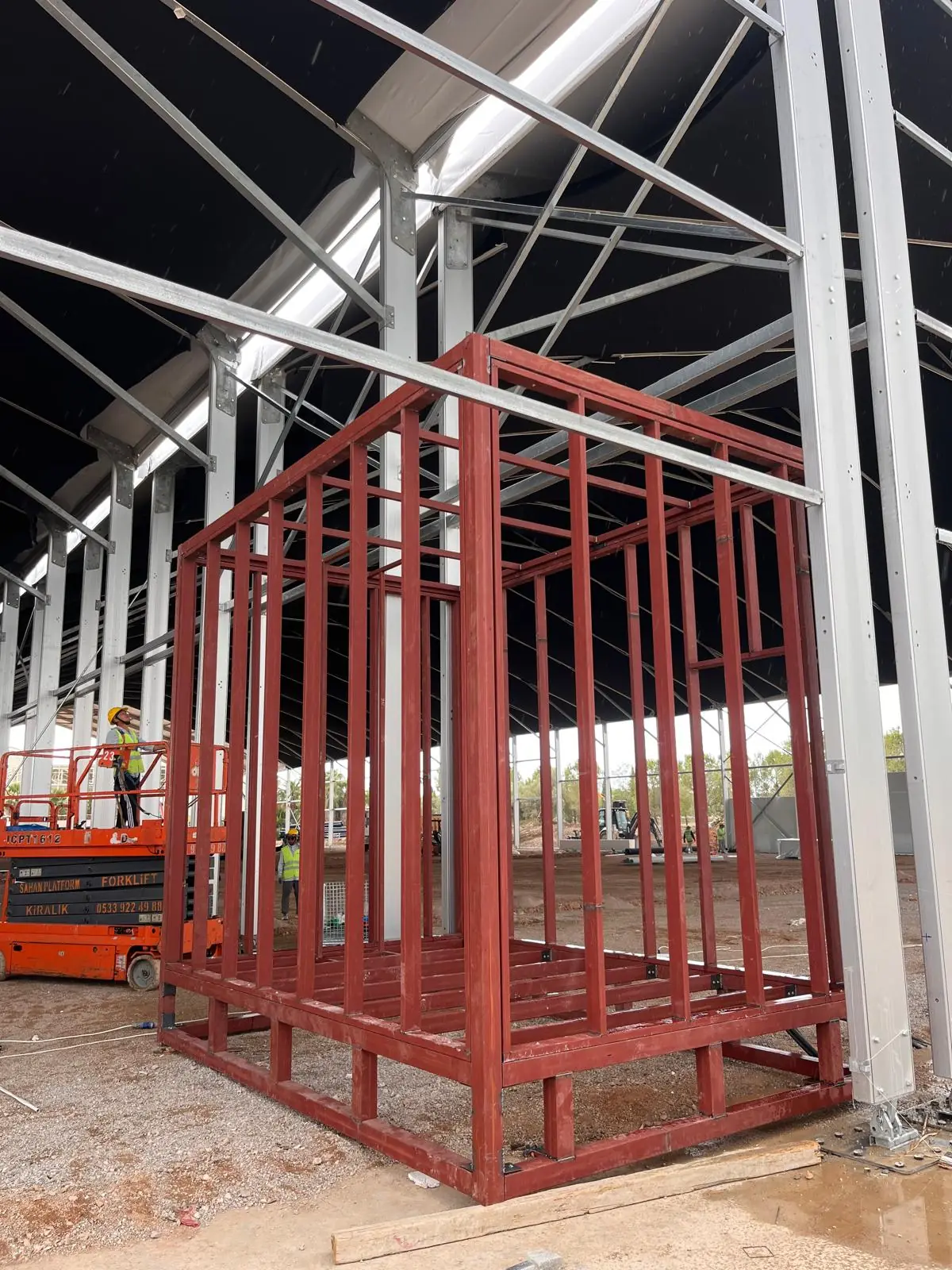 A red-primed steel cage module inside a large hangar.