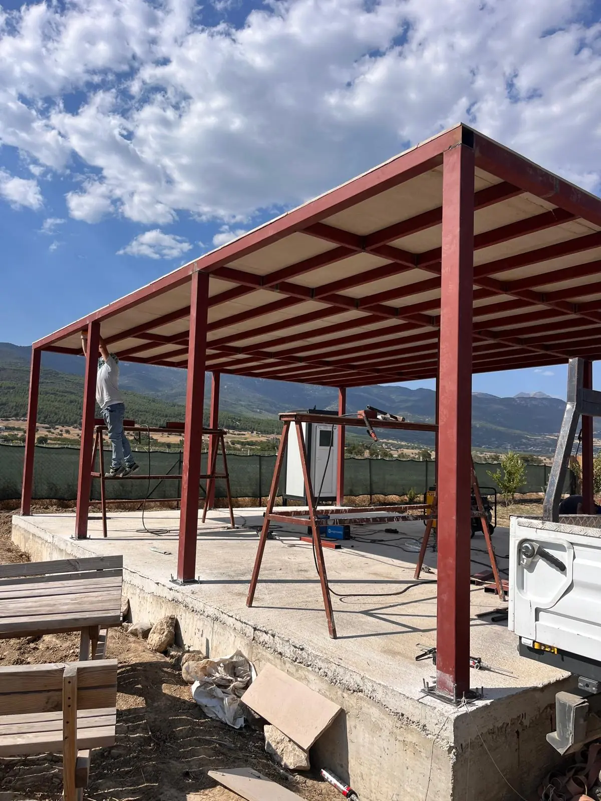 Red steel canopy in front of mountains, on a concrete base with timber benches.